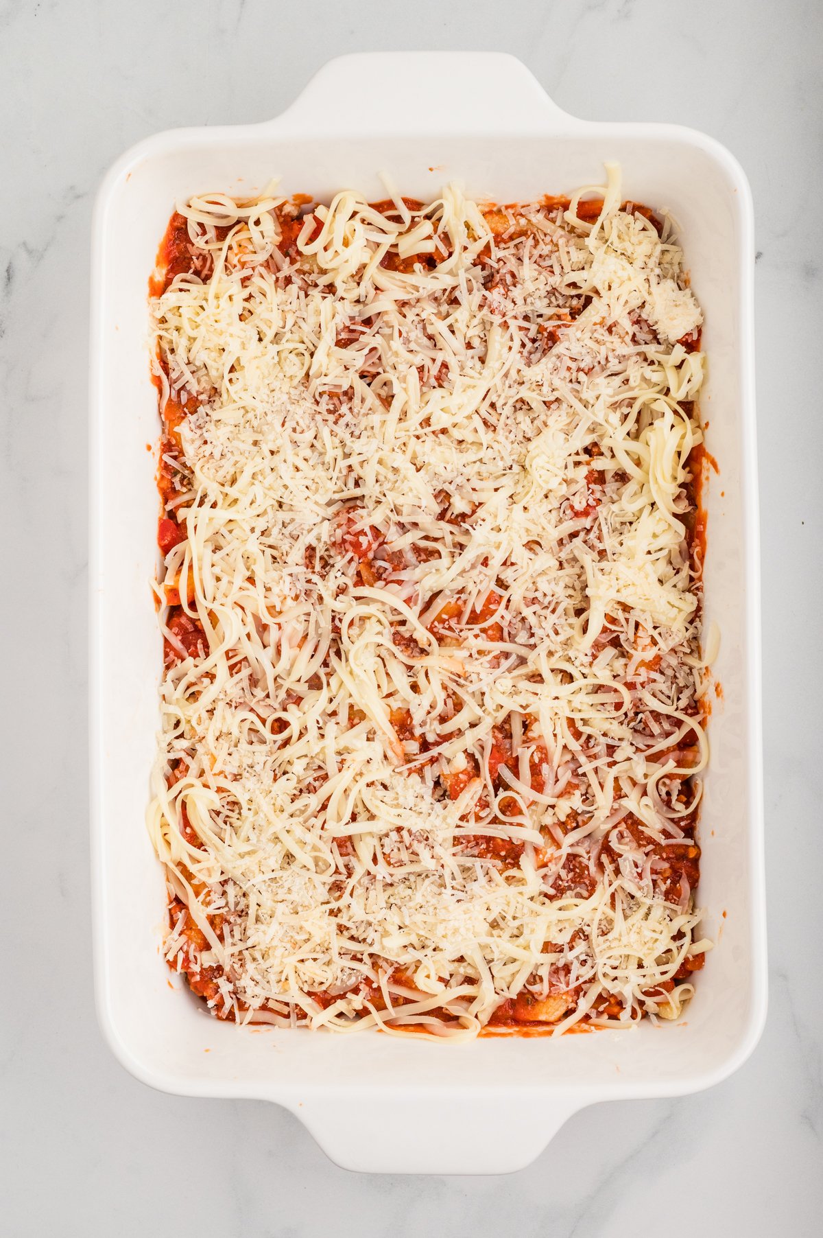 Top-down image of the white casserole pan with shredded mozzarella and grated Parmesan sprinkled over the final sauce-and-pasta layer, ready for baking.