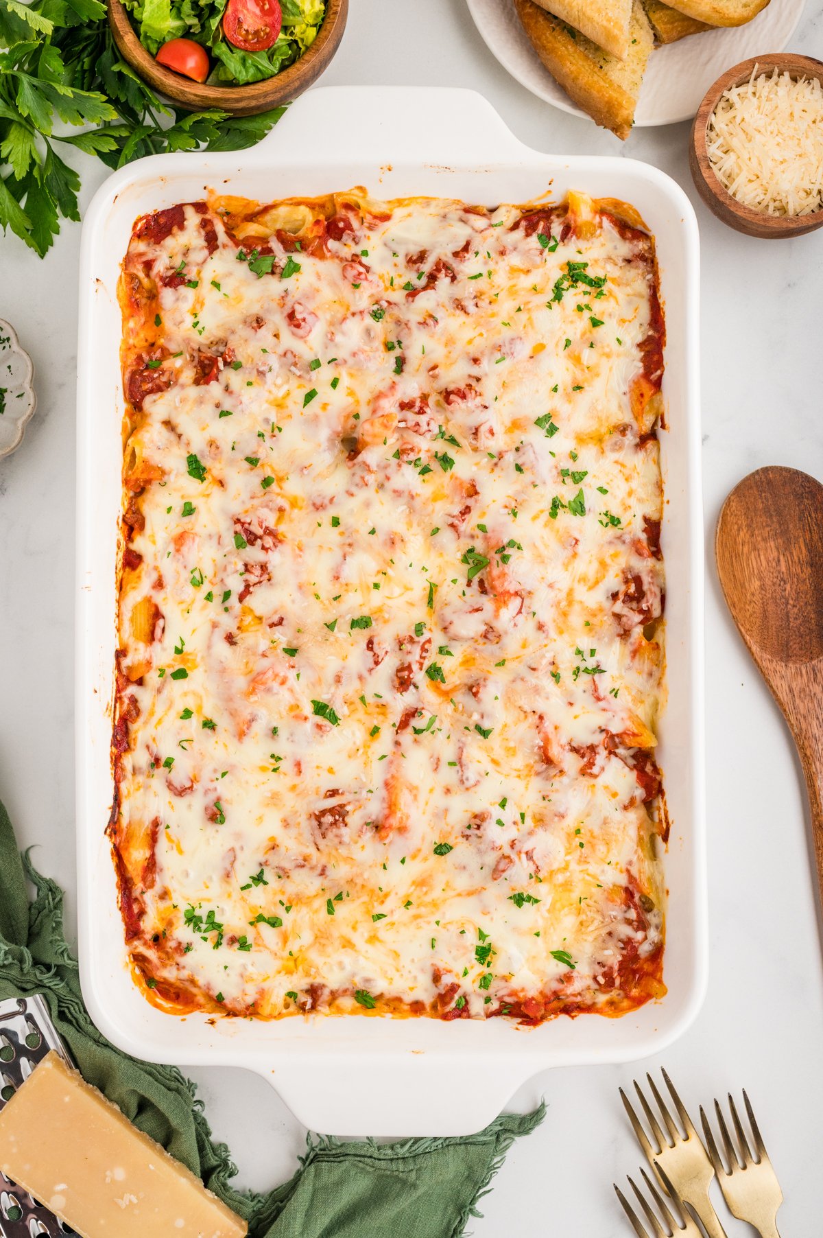 Overhead shot of a fully baked Lazy Lasagna in a white baking dish, topped with melted, golden cheese and sprinkled with chopped parsley. Surrounding the dish are bowls of fresh salad, breadsticks, and a small bowl of grated Parmesan.