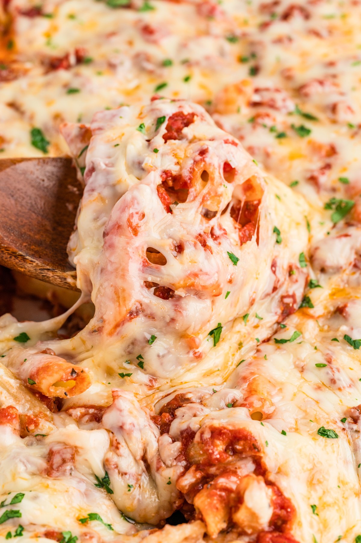 Close-up of a wooden spatula lifting a portion of cheesy Lazy Lasagna from a white baking dish, showing melted mozzarella stretching over penne pasta and red tomato-meat sauce, sprinkled with chopped parsley.