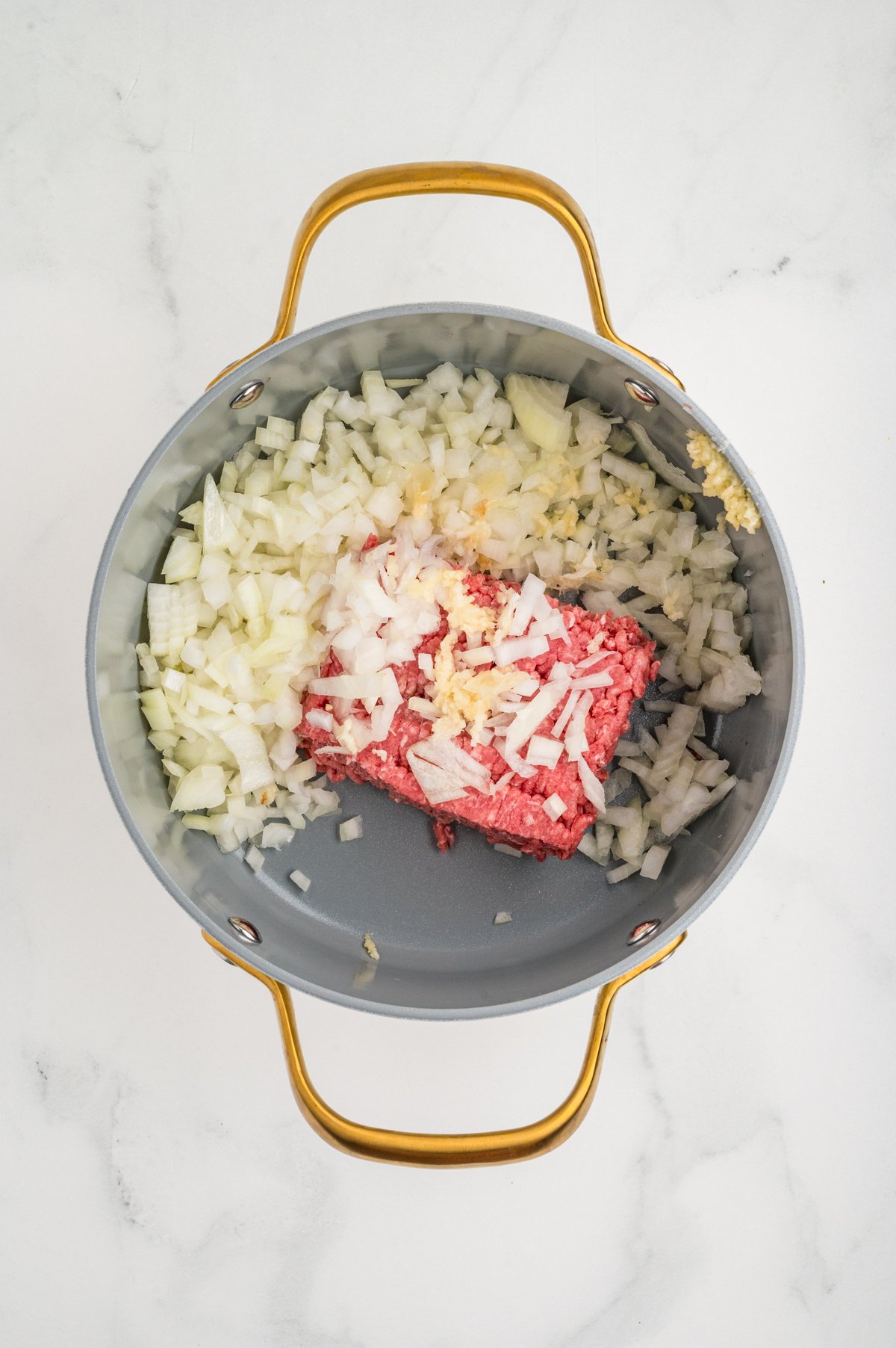 Overhead shot of a light-gray saucepan holding raw ground beef in the center, surrounded by a mound of diced white onion and minced garlic.