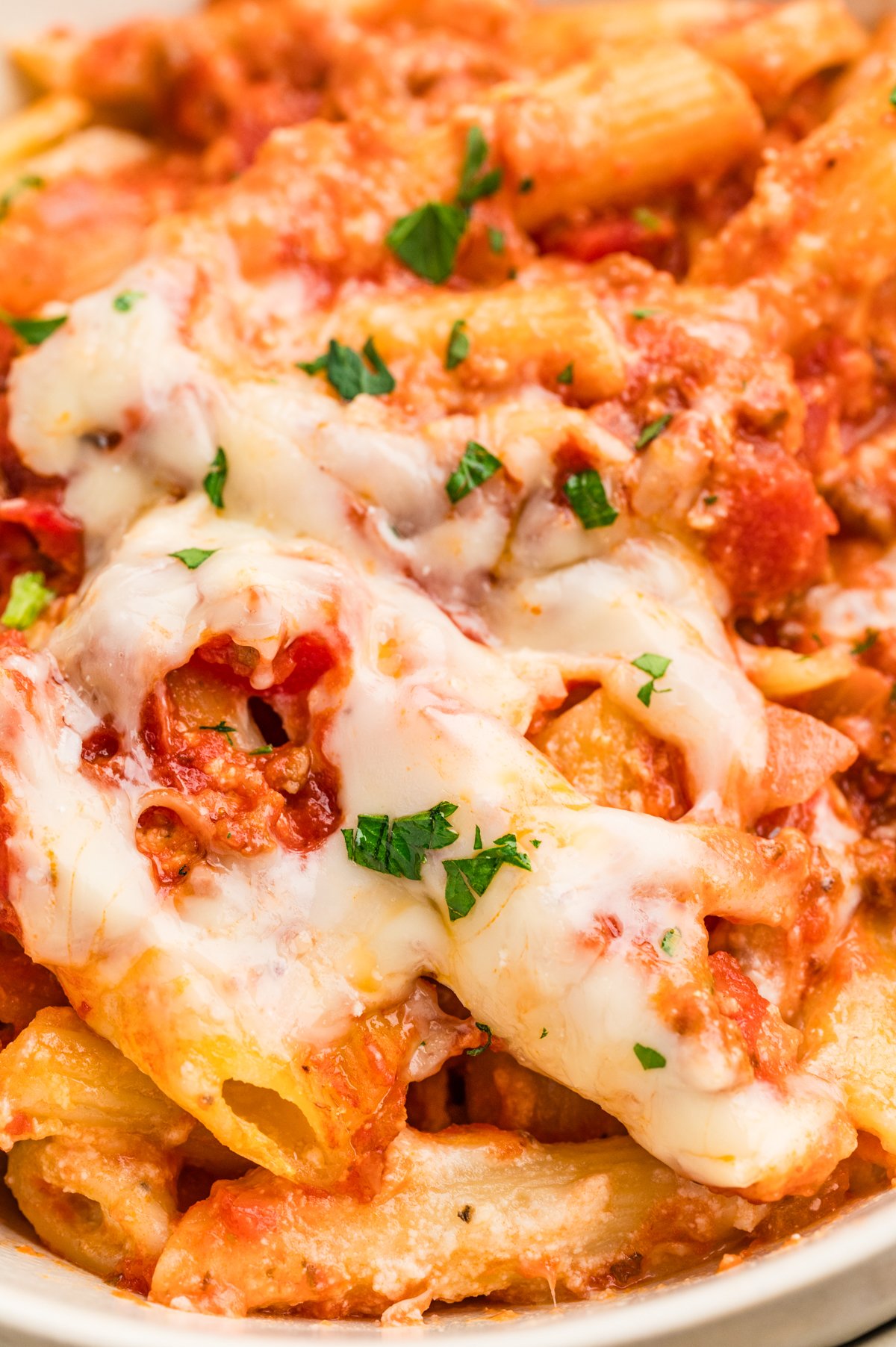 Extreme close-up of cheesy Lazy Lasagna in a white bowl, with gooey melted mozzarella over penne and tomato-beef sauce, and bits of parsley on top.