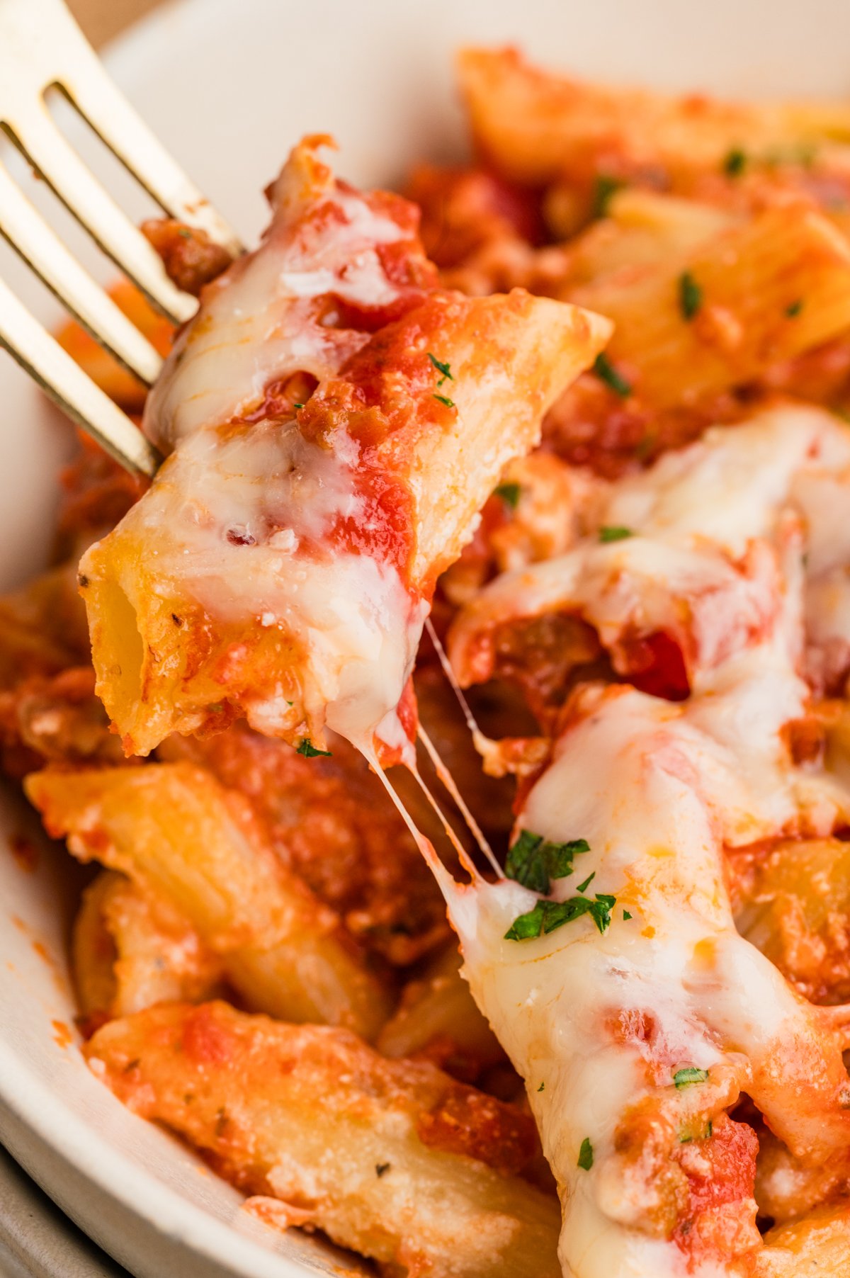 Overhead shot of a white bowl filled with Lazy Lasagna, featuring penne coated in tomato-meat sauce and a layer of melted cheese, lightly garnished with parsley on a fork.