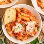 Top-down view of three white bowls of Lazy Lasagna on a woven placemat, each serving topped with melted cheese and parsley; the front bowl also includes a piece of garlic bread resting on the lasagna.
