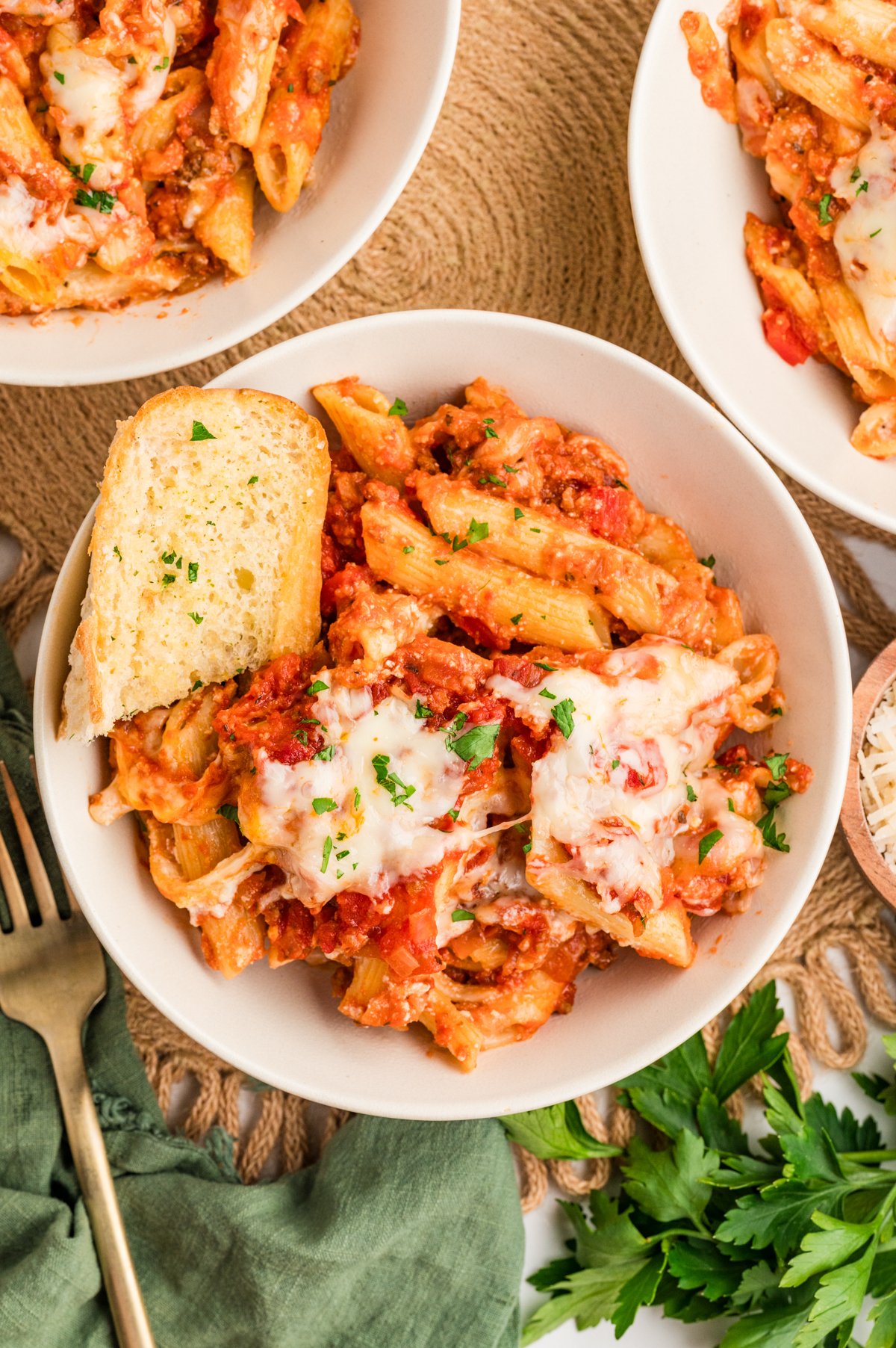 Top-down view of three white bowls of Lazy Lasagna on a woven placemat, each serving topped with melted cheese and parsley; the front bowl also includes a piece of garlic bread resting on the lasagna.