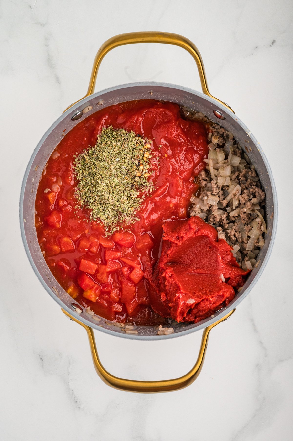 iew of a saucepan filled with browned ground beef and onions on one side, and on top of that a spoonful of tomato paste, a pile of dried Italian seasoning, diced tomatoes, and crushed tomato sauce ready to be stirred.