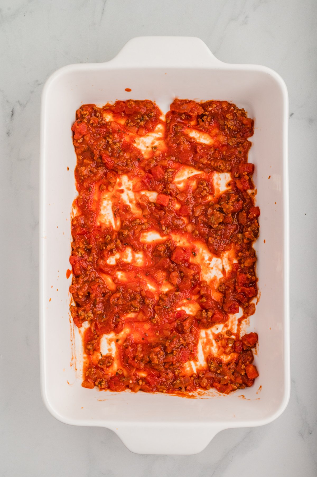 Top view of an empty white 9×13 baking dish with about half a cup of meat-tomato sauce spread thinly across the bottom.