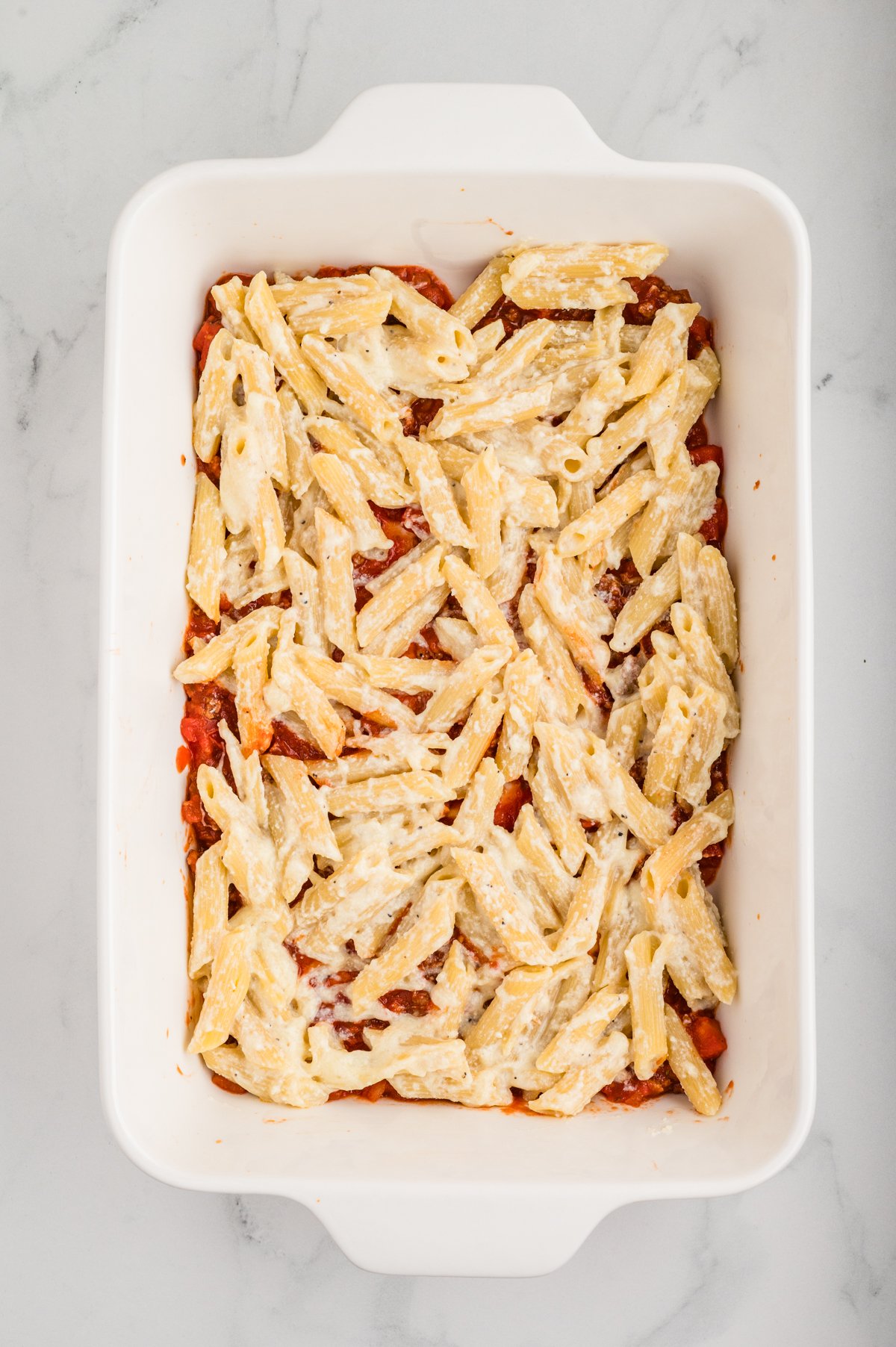 Overhead shot of a white casserole dish showing half of the pasta-cheese mixture (penne coated in ricotta-cheese) layered on top of the initial sauce.