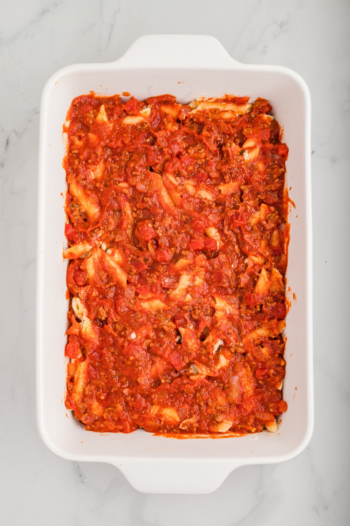 View of the same white baking dish now with the remaining meat-tomato sauce spread evenly over the cheese-pasta layer.