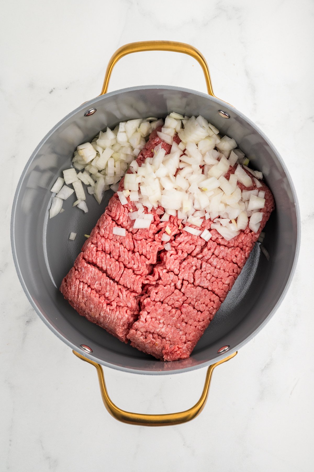 Top-down view of a large skillet containing raw lean ground beef and diced white onion over a white countertop.
