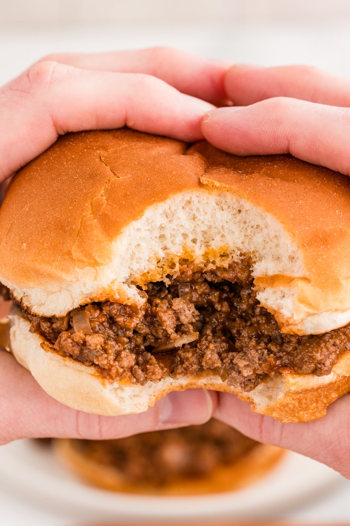 Close-up of two hands holding a barbecue ground beef sandwich in a soft hamburger bun, with a bite taken out of it, revealing saucy seasoned beef inside.