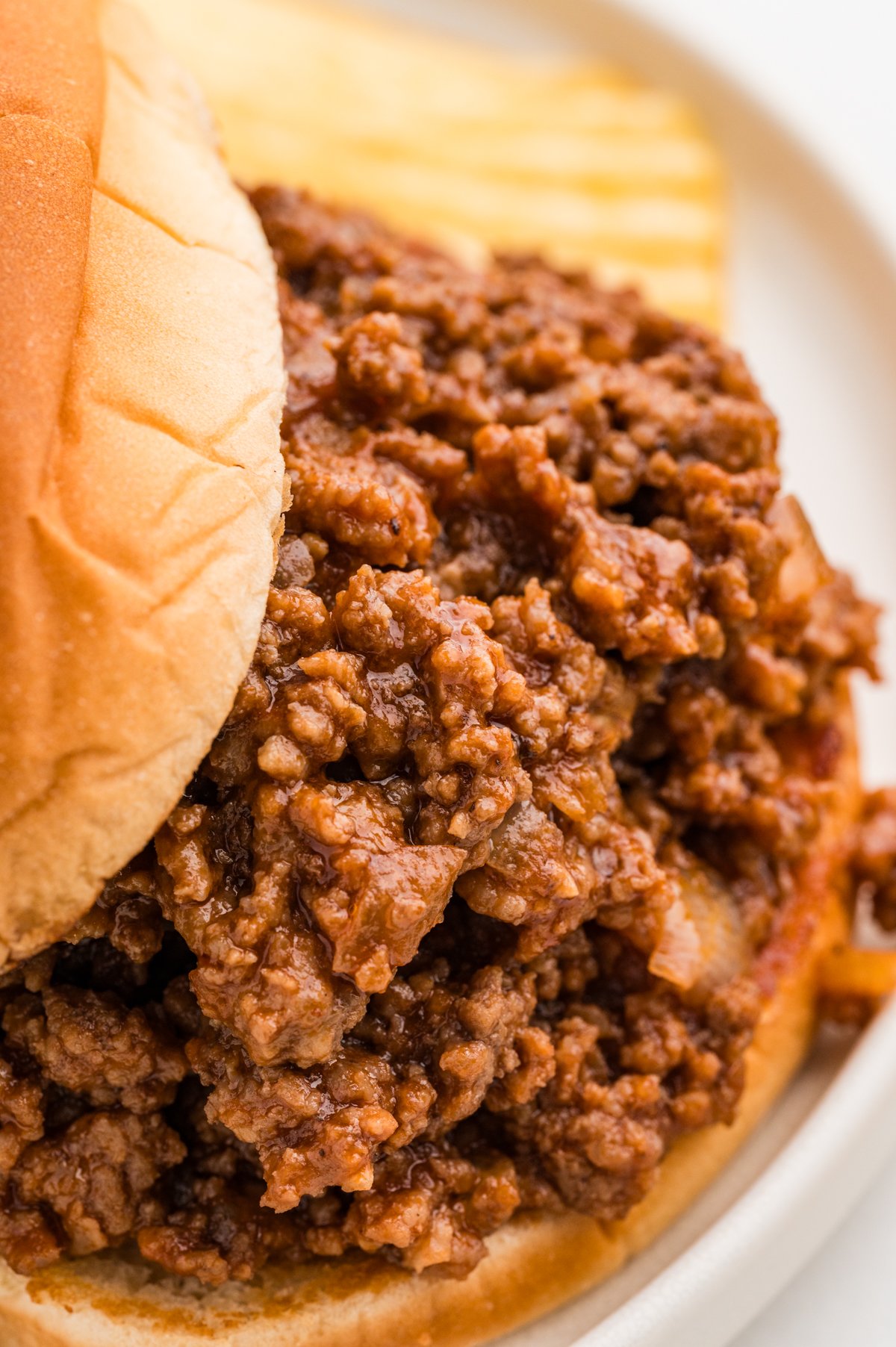 Close-up of saucy BBQ ground beef filling spilling out of a soft hamburger bun, with ridged potato chips in the blurred background.