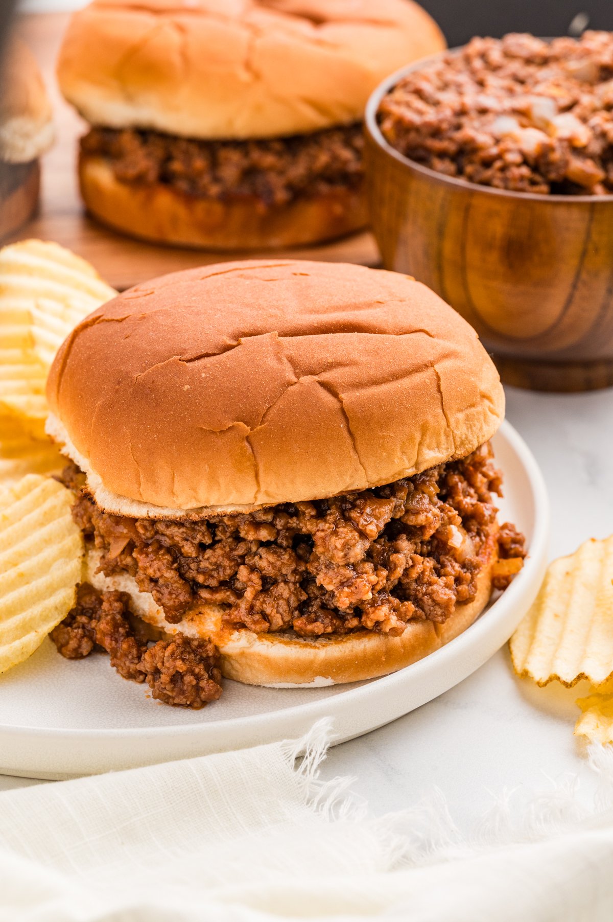 Front-on shot of three barbecue ground beef sandwiches lined up on a wooden serving board, with the middle sandwich in clear focus and a bowl of extra beef mixture blurred in the background.