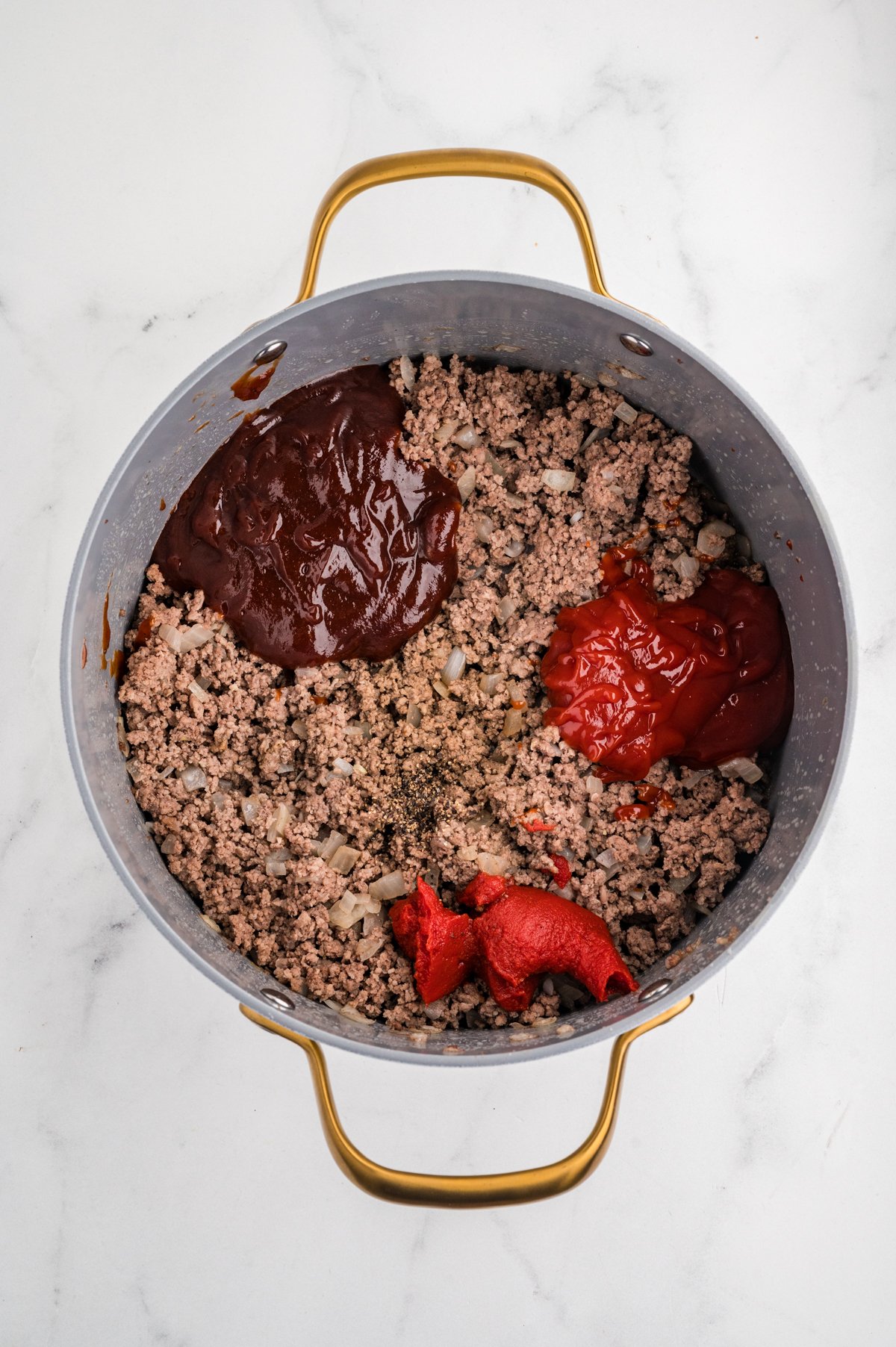 Top-down view of cooked ground beef and onion in a skillet with separate dollops of barbecue sauce, ketchup, and tomato paste ready to be stirred in.