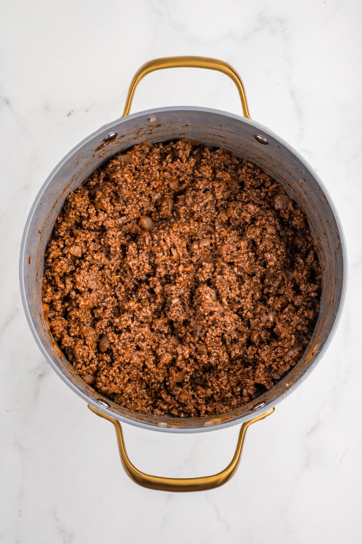 Top-down view of a large skillet containing cooked ground beef and onion over a white countertop.