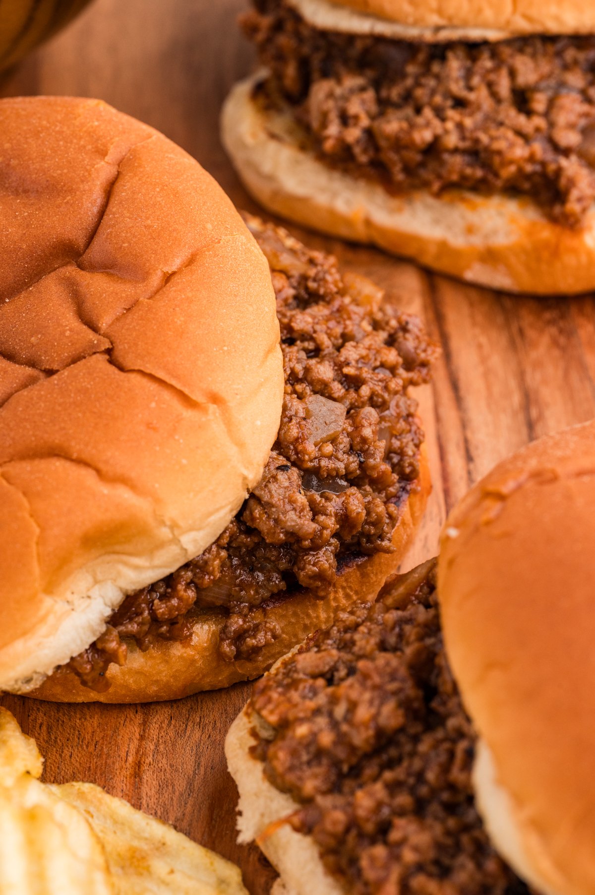 Tight close-up of saucy barbecue ground beef filling spilling out of a soft hamburger bun, with ridged potato chips in the blurred background.
