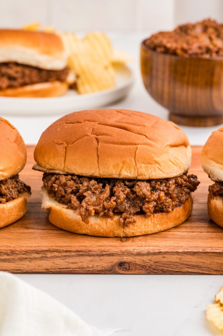 Angled close-up of three barbecue ground beef sandwiches on a wooden board, showing saucy beef spilling onto the bun edges and a handful of potato chips in the foreground.