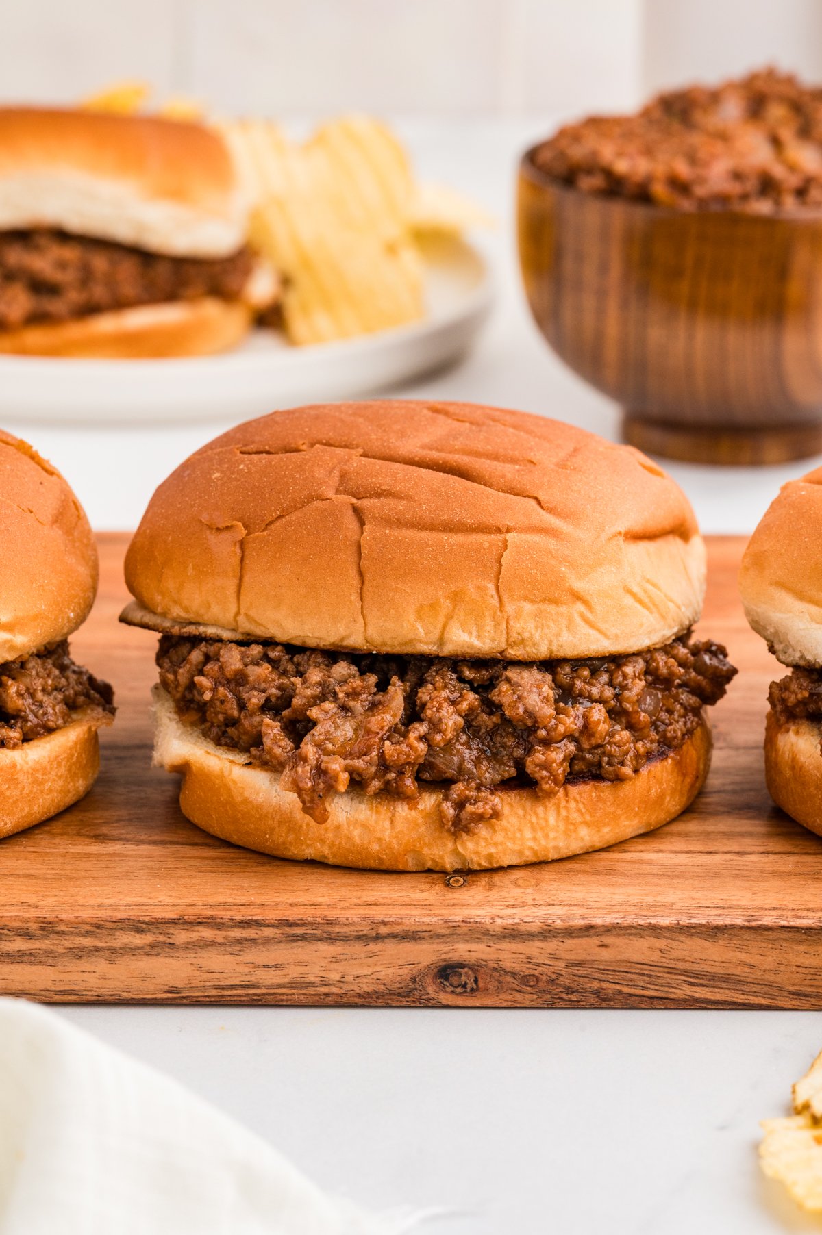 Angled close-up of three barbecue ground beef sandwiches on a wooden board, showing saucy beef spilling onto the bun edges and a handful of potato chips in the foreground.