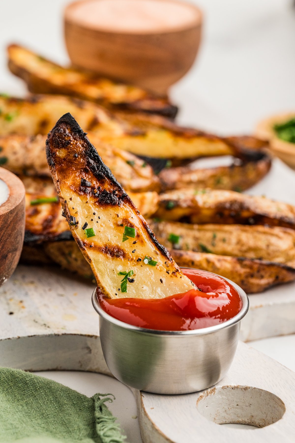 Portrait-oriented close-up of a single grilled potato wedge held upright and dipped into a small metal cup of ketchup.