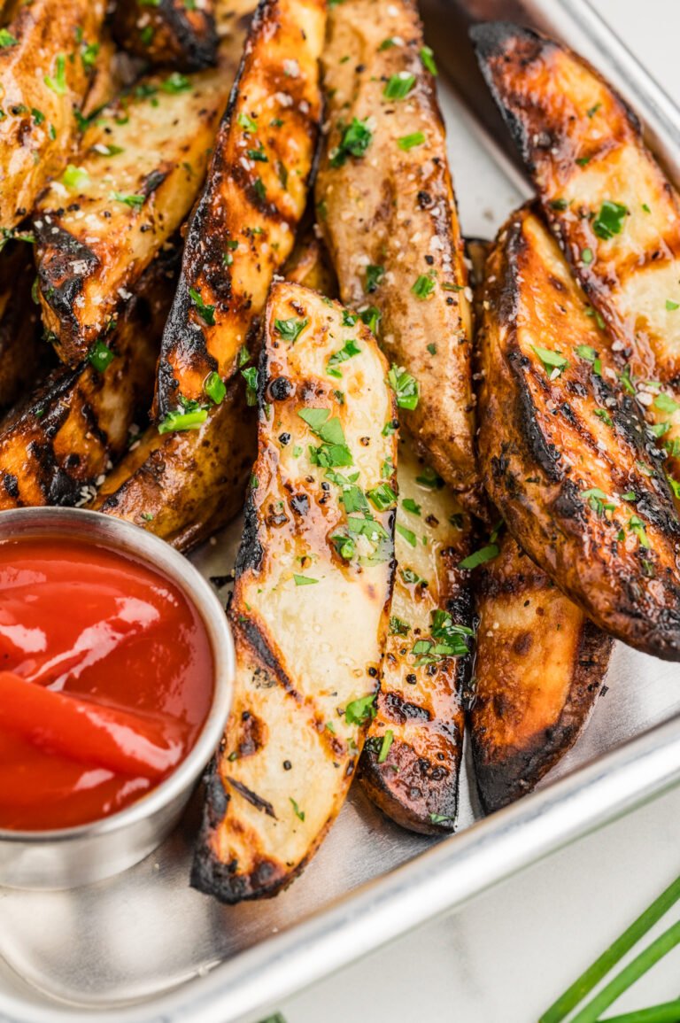 Tight close-up of a stack of grilled potato wedges in a metal tray, each wedge coated with melted herb butter, black pepper, and chopped parsley. A small metal cup of ketchup appears at bottom left.
