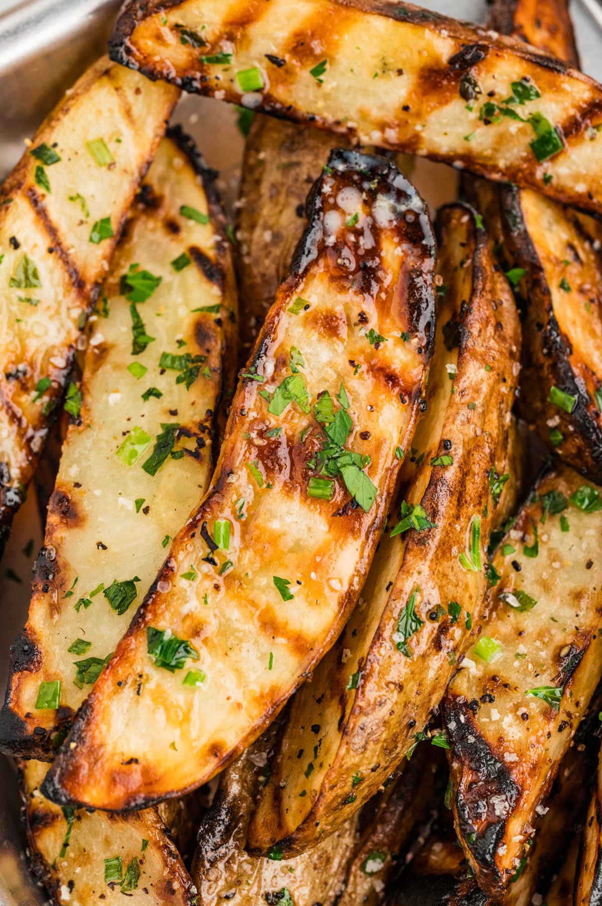 Close-up overhead of several grilled potato wedges crowded together in a metal tray, showing golden interiors, dark grill marks, and flecks of chopped parsley and chives.