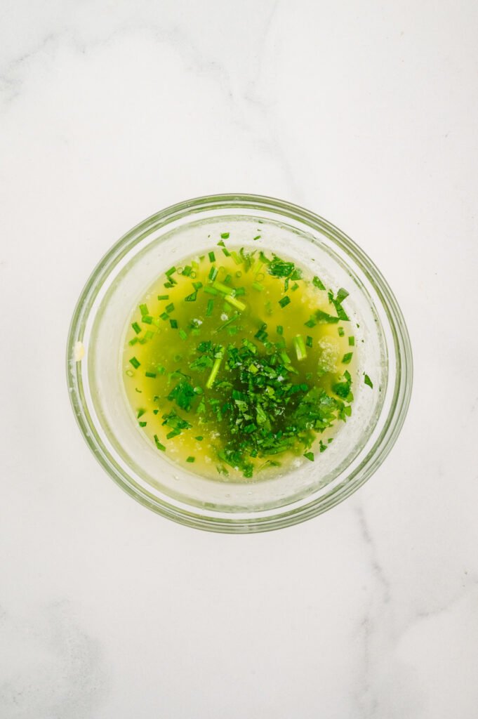View of a small glass mixing bowl containing melted butter whisked together with finely chopped parsley and chives.