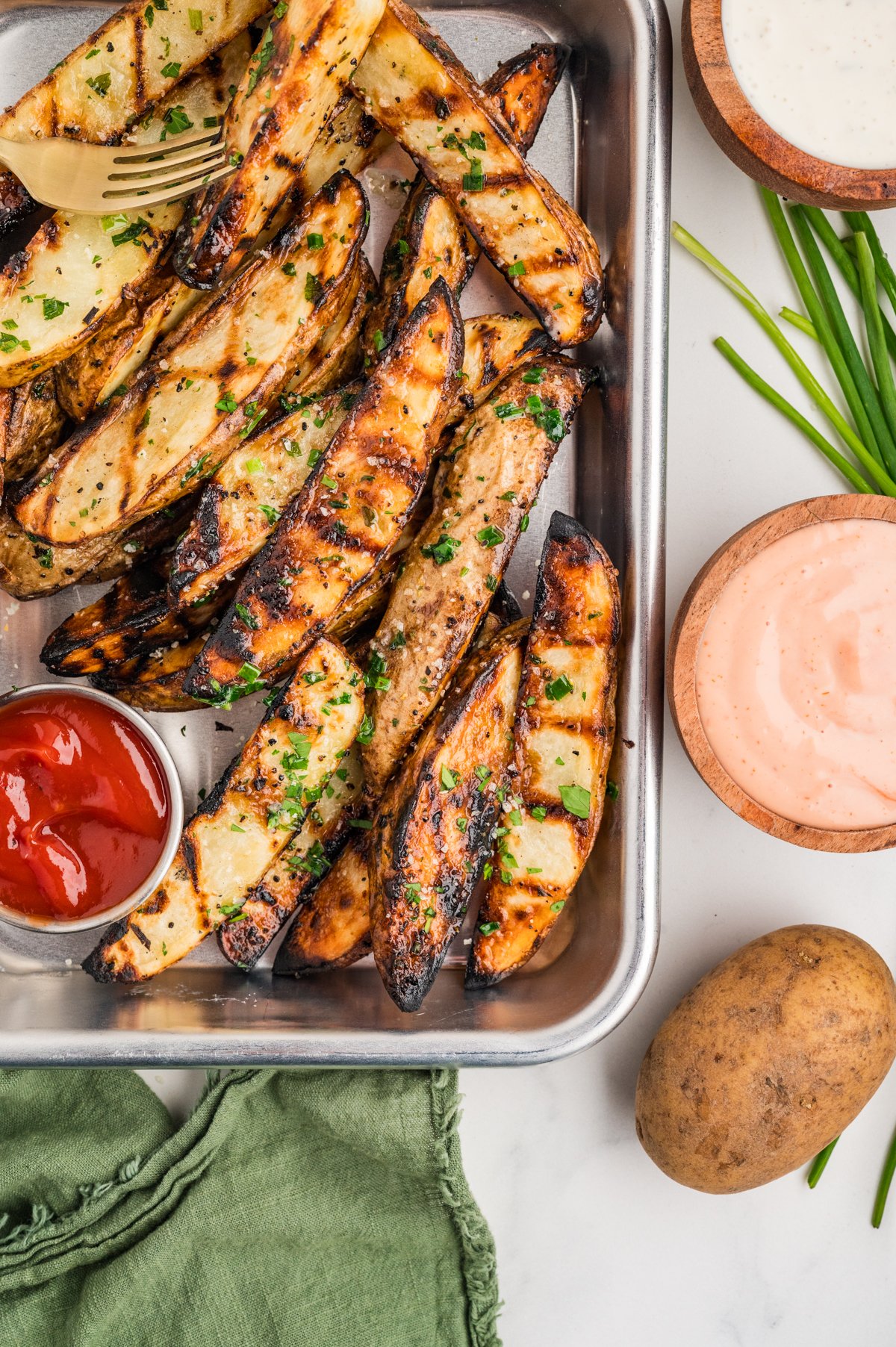 Overhead shot of a metal baking tray filled with grilled potato wedges, each marked with charred grill lines and sprinkled with parsley and chives.