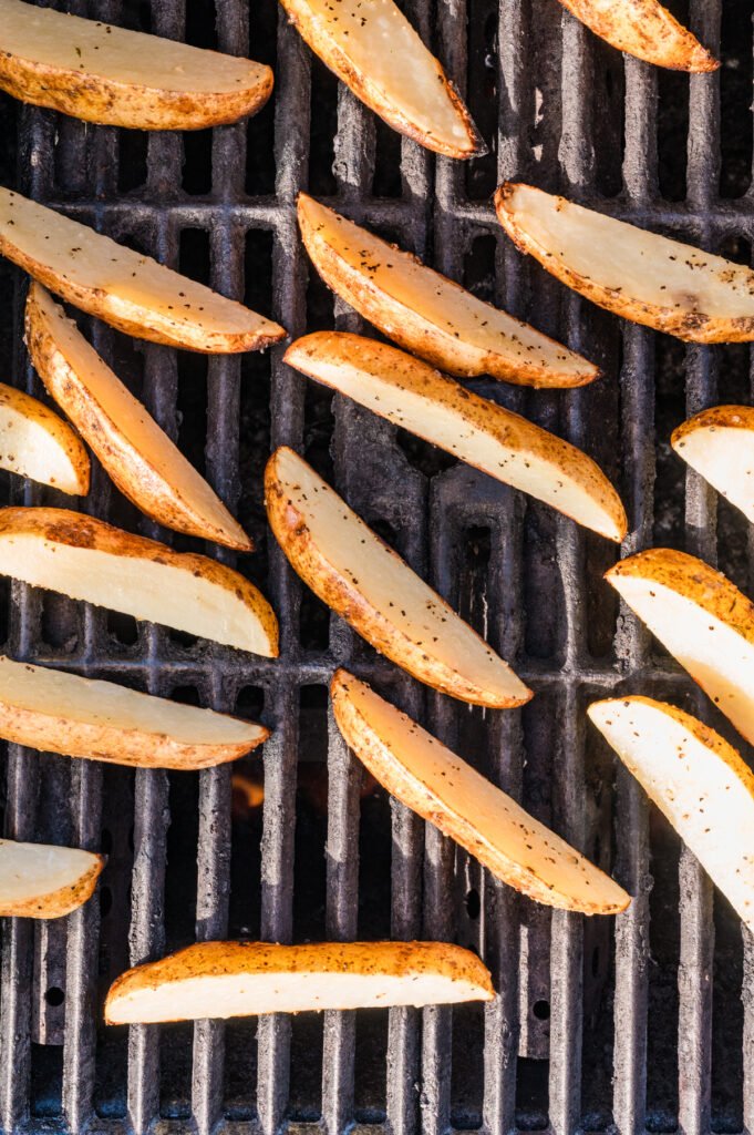 Close-up overhead shot of potato wedges placed directly on hot grill grates, just beginning to char and cook.