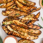 Overhead view of a white wooden cutting board piled with grilled potato wedges garnished with chopped parsley and chives.