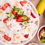 A top-down view of the finished strawberry banana cheesecake salad in a white serving bowl, garnished with strawberry slices, banana rounds, and small strawberry blossoms, surrounded by additional fruit on a pink marble background.