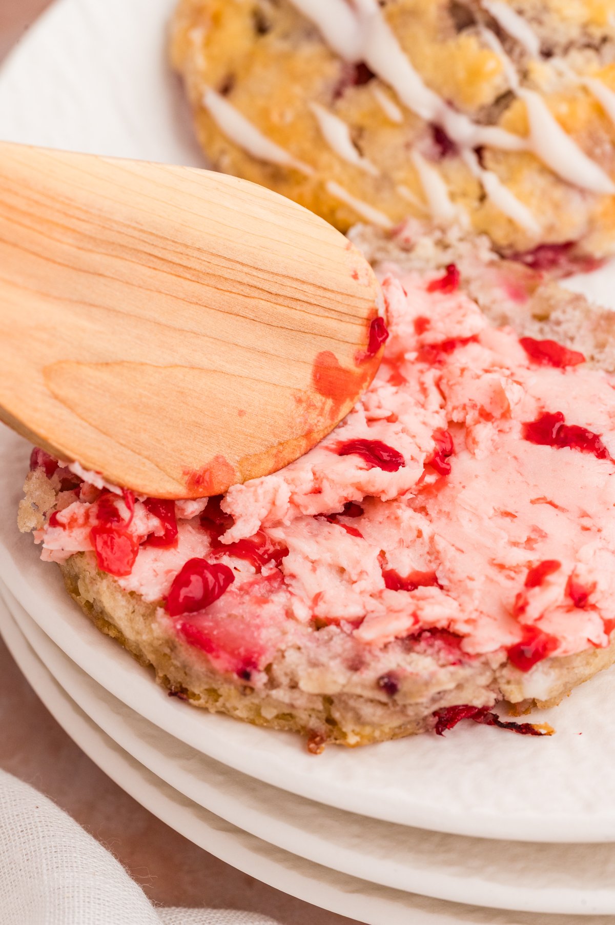 A halved biscuit on a white plate being spread with pink strawberry butter using a wooden spatula.