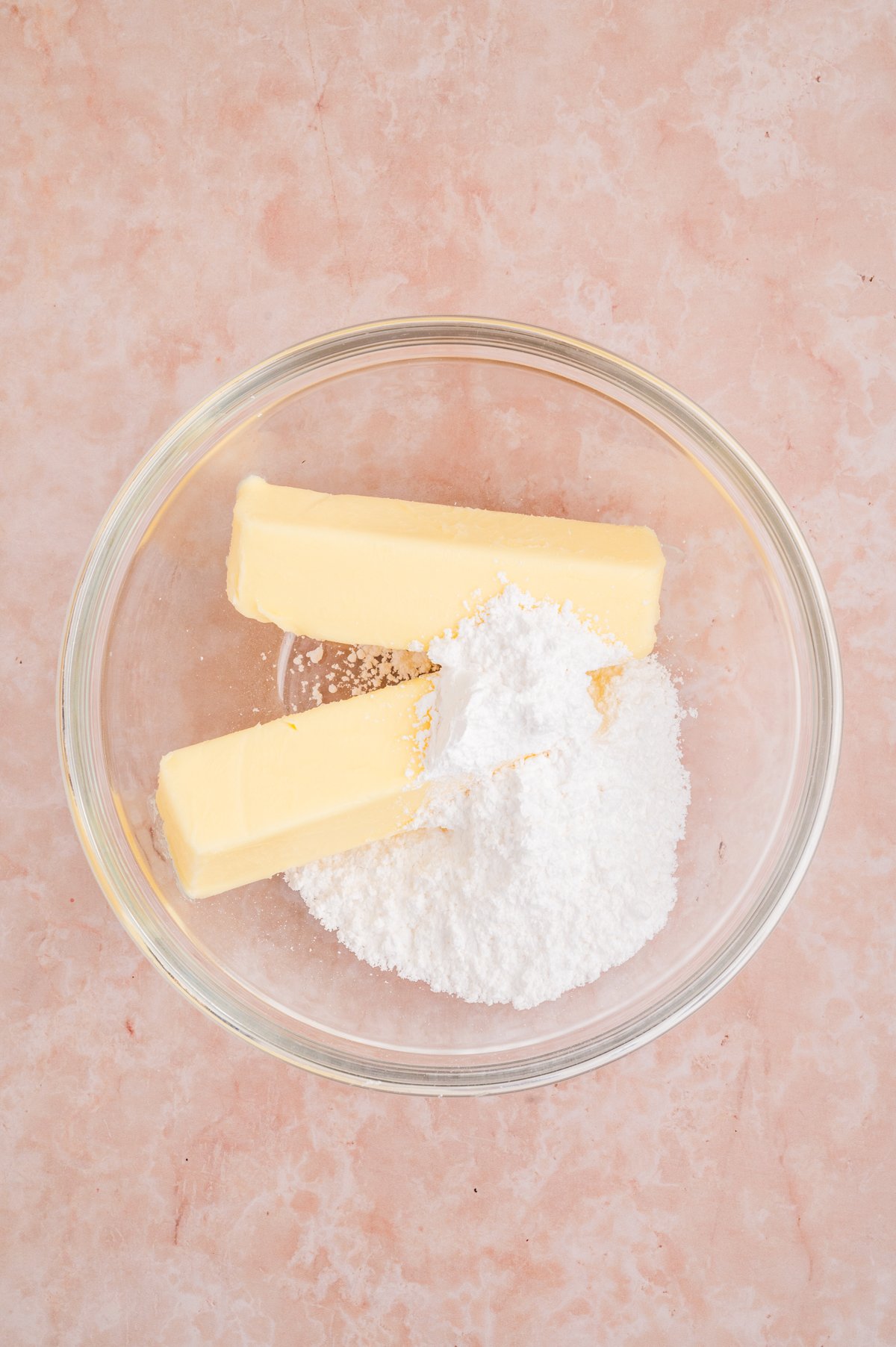 Two sticks of softened butter and a mound of powdered sugar are in a clear bowl.