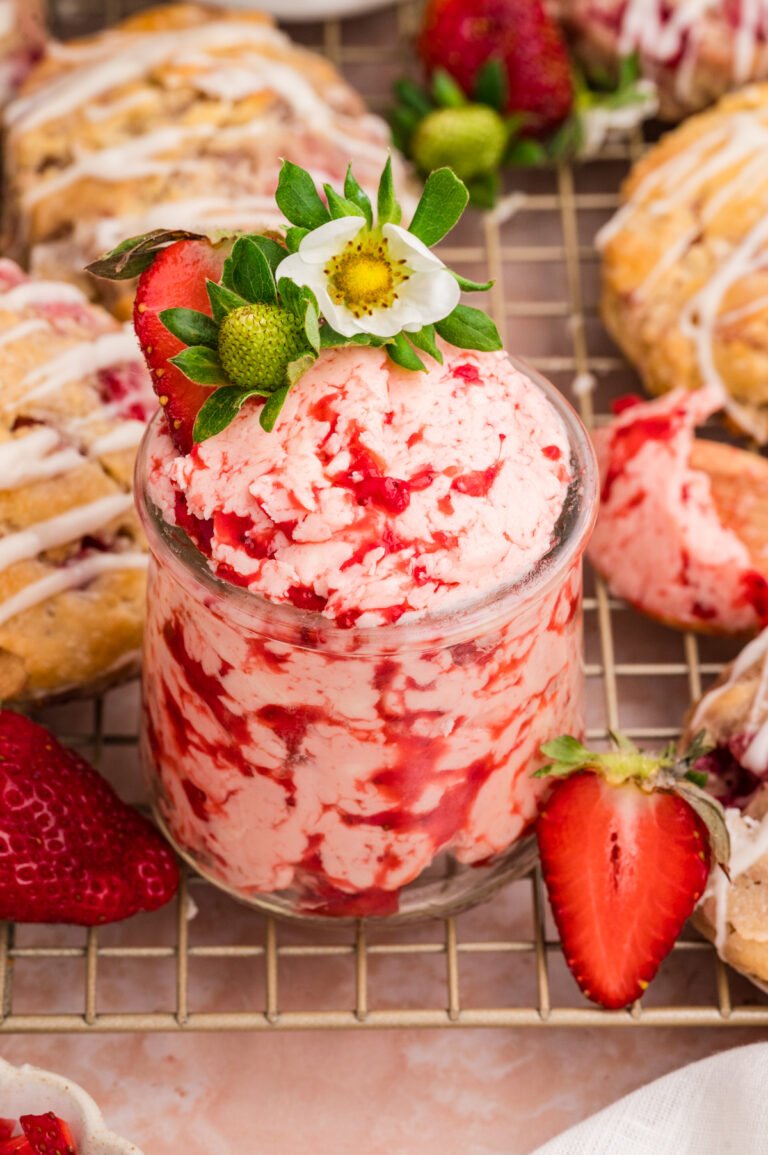 A small glass jar filled with pink strawberry butter, surrounded by fresh strawberries and drizzled biscuits on a cooling rack.