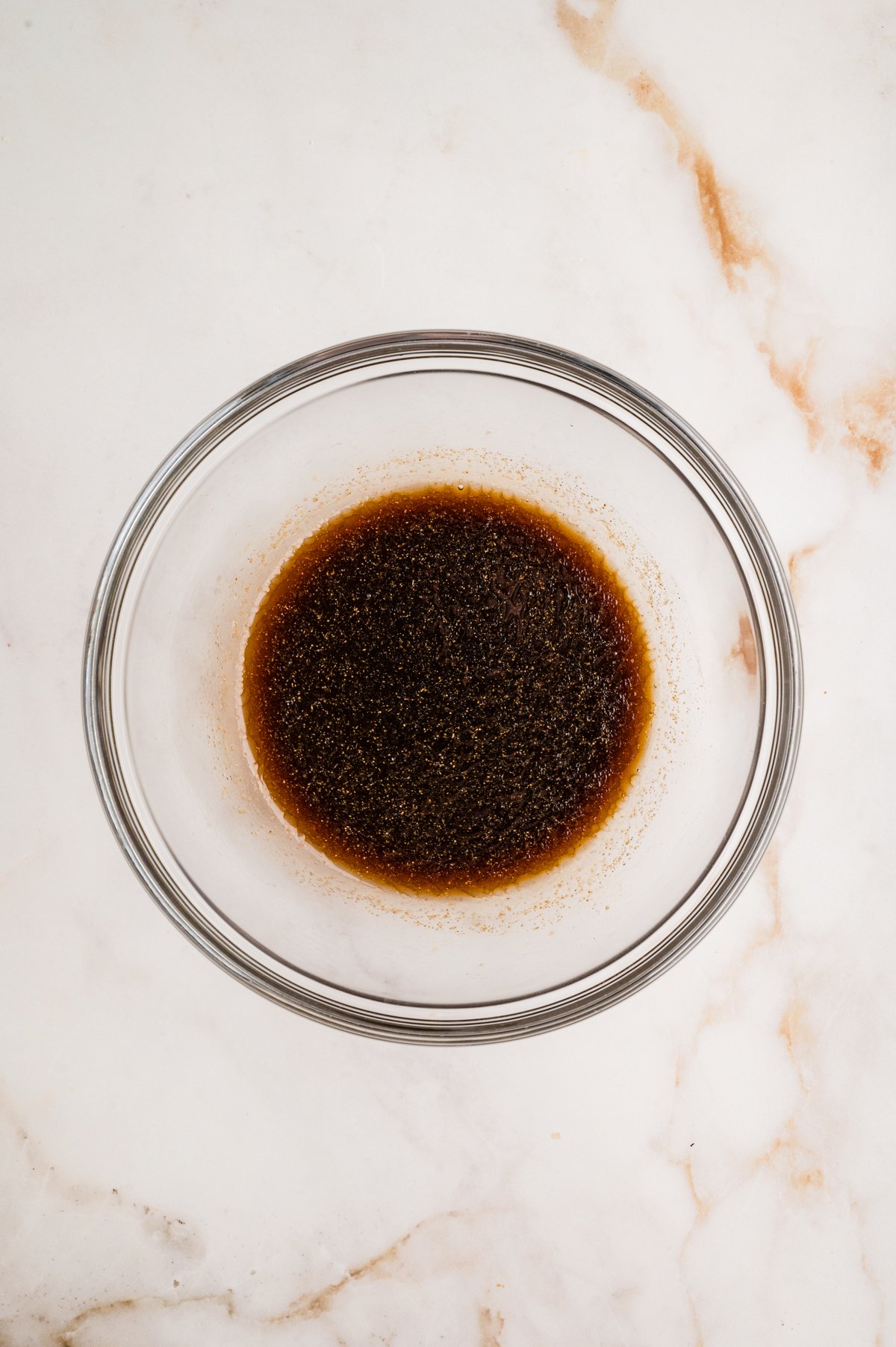 A clear glass bowl sitting on a marble surface filled with dark brown marinade.