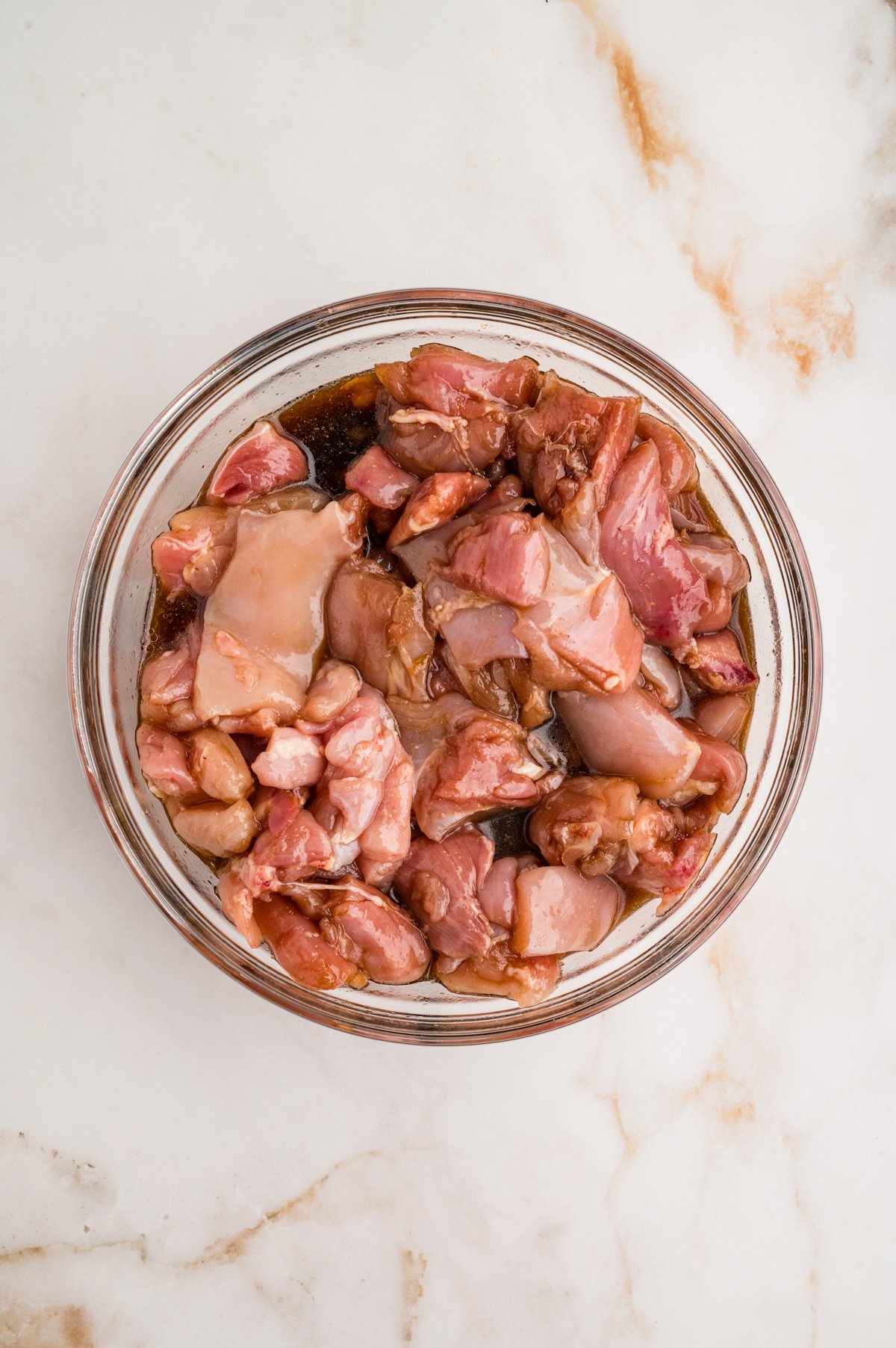 Glass mixing bowl with bite-sized raw chicken thighs soaking in marinade.