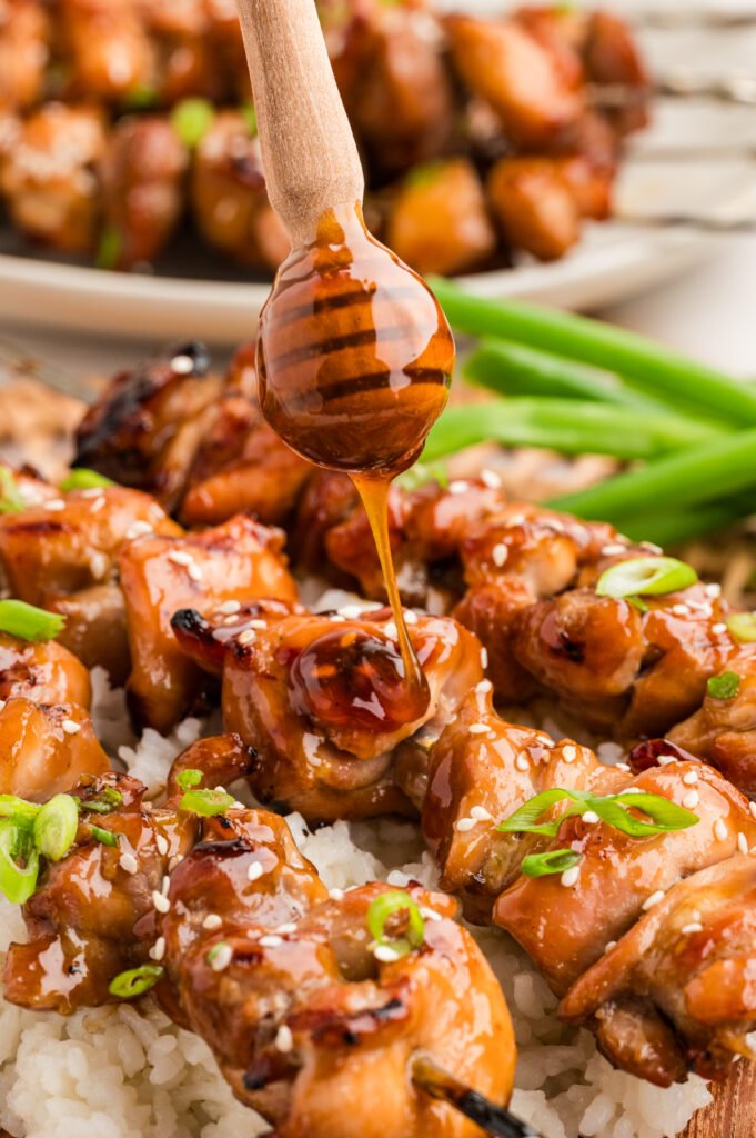 Teriyaki sauce being drizzled from a wooden honey dipper onto grilled chicken skewers resting on rice.