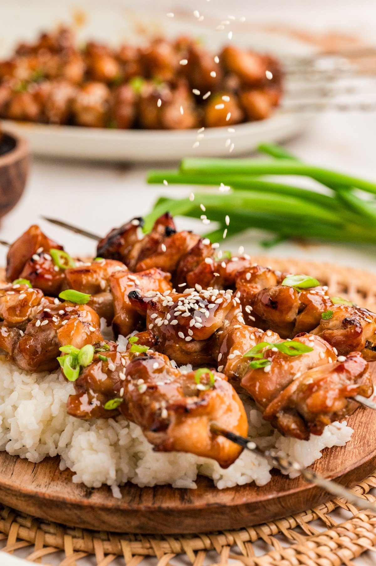Action shot of sesame seeds falling onto glazed chicken skewers on a bed of rice, with green onions in the background.