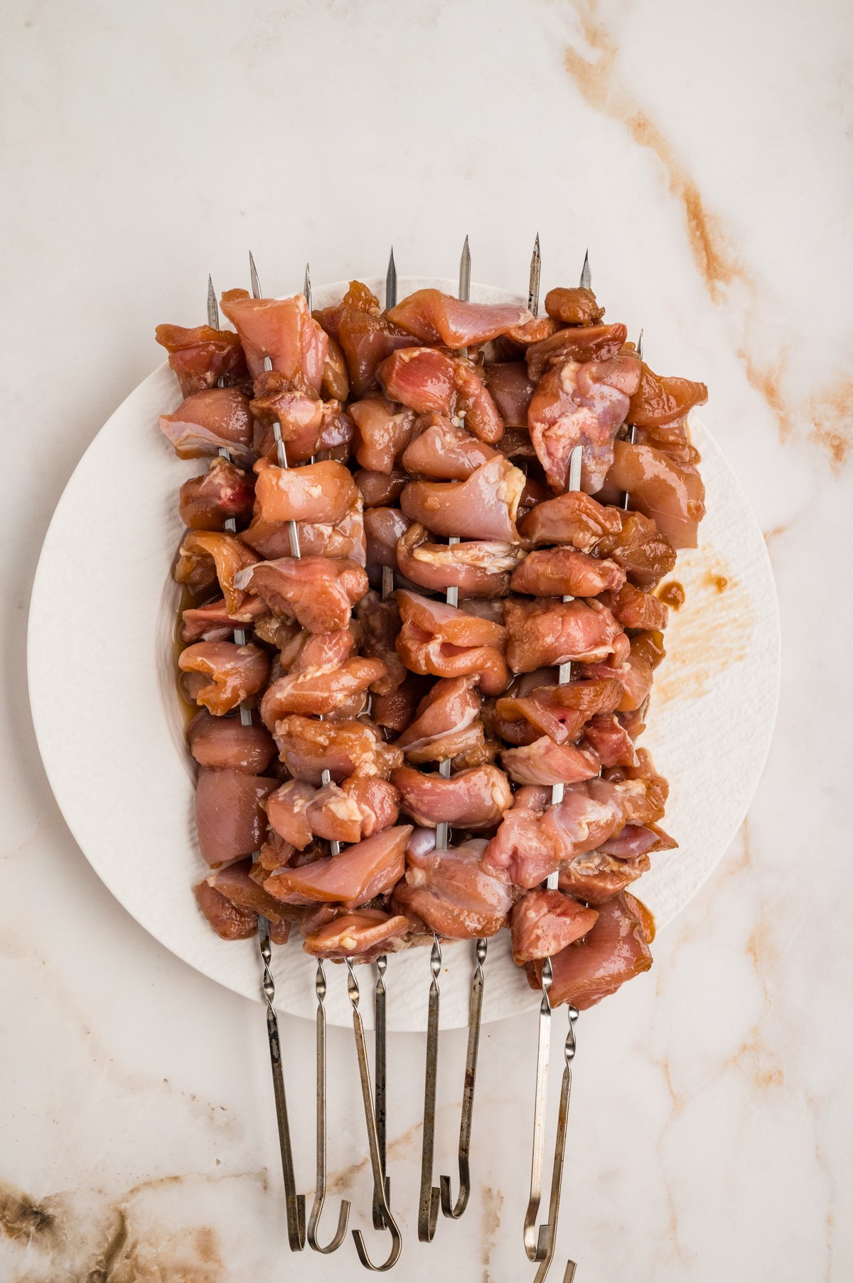 Metal skewers threaded with marinated chicken pieces arranged on a white plate.