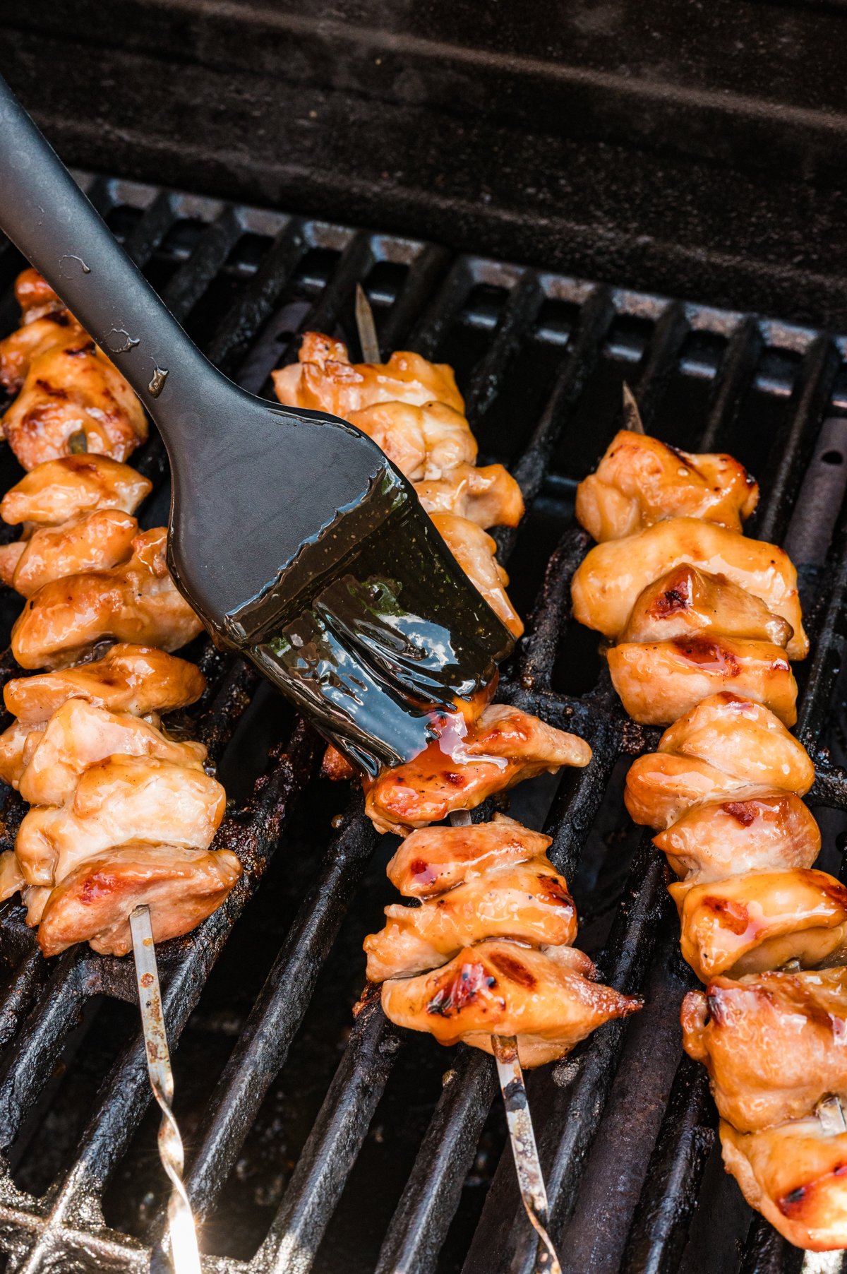 A brush applying glossy teriyaki sauce onto chicken skewers as they cook on the grill.