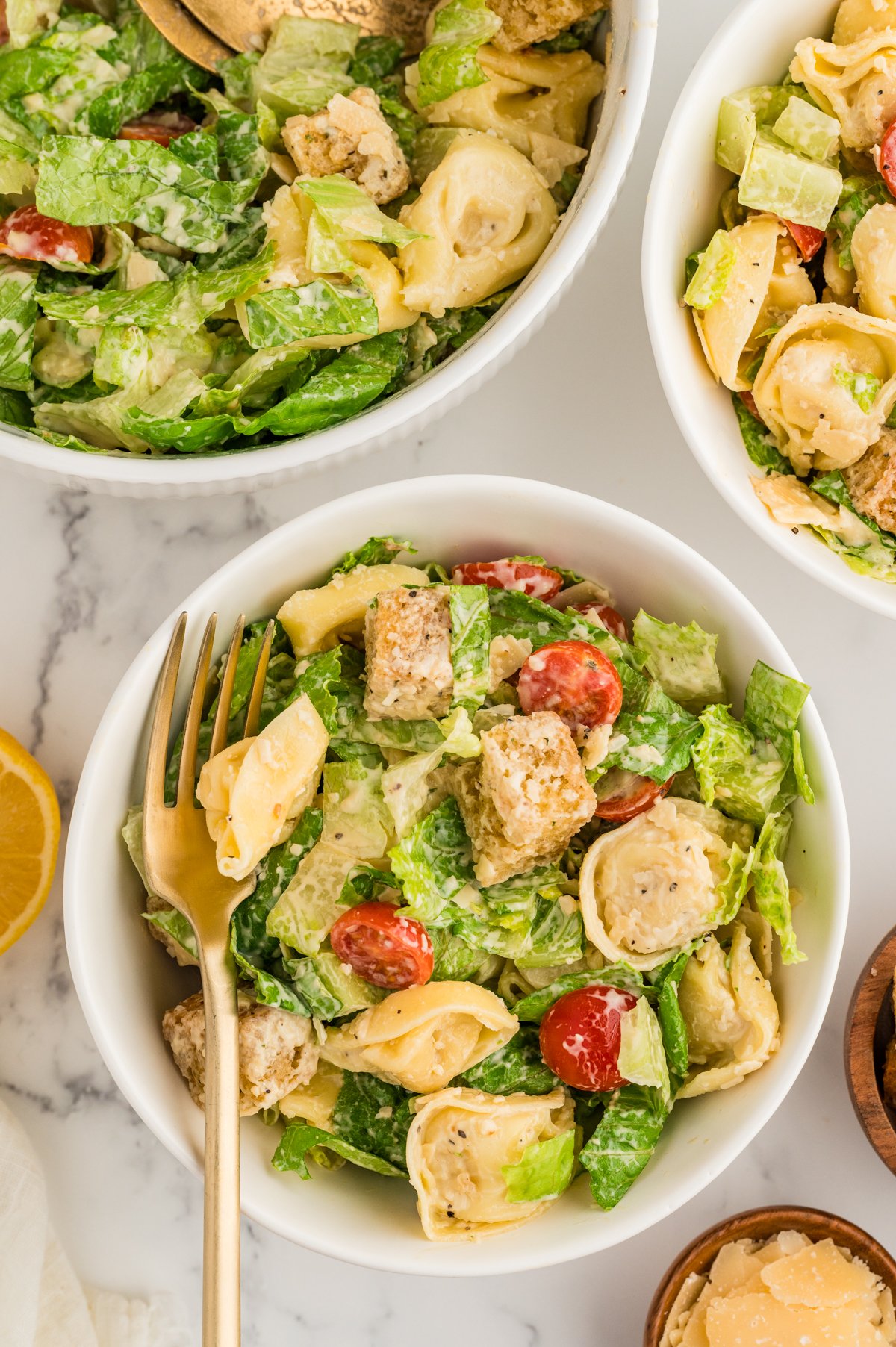 Individual servings of tortellini Caesar salad in white bowls with gold fork and visible lettuce and tomatoes.