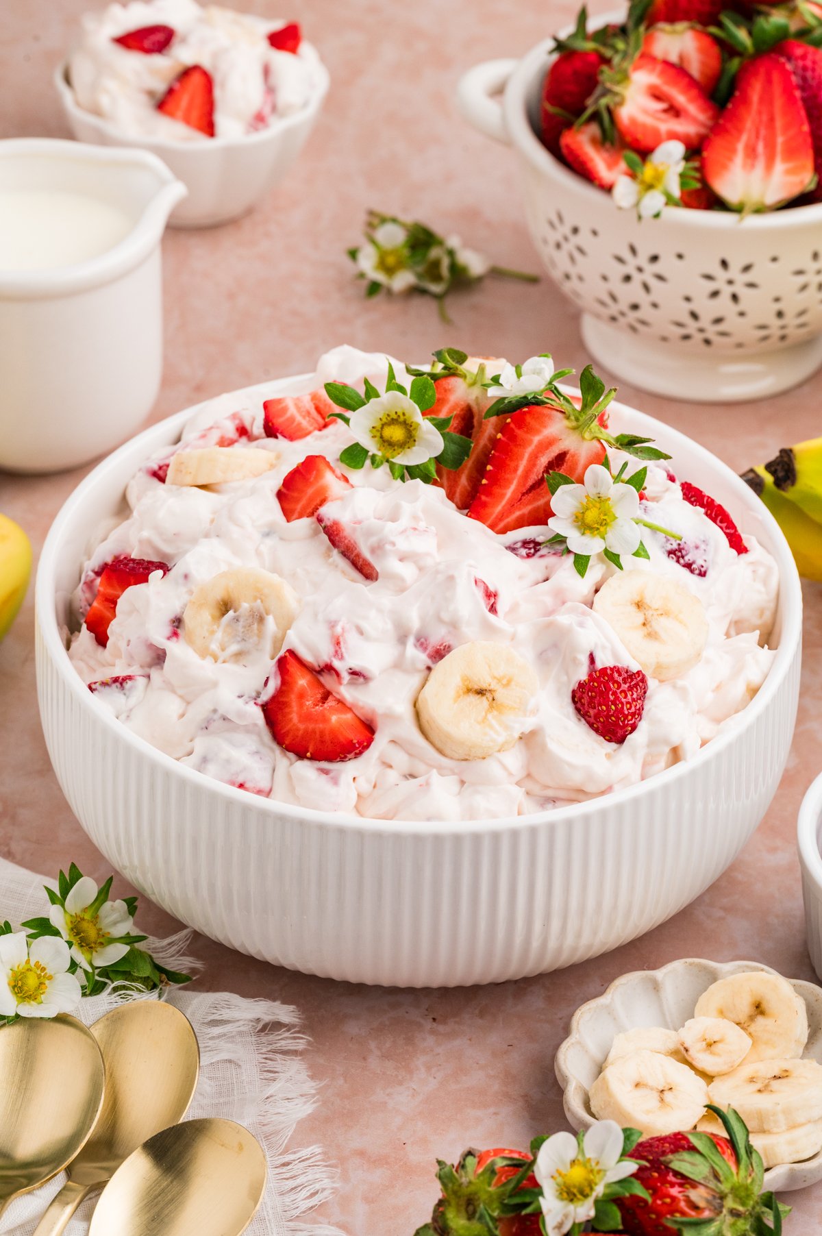 A white ribbed serving bowl filled with creamy pink cheesecake salad mixed with sliced strawberries and bananas, garnished with halved strawberries and small white strawberry blossoms, surrounded by fresh fruit, a small pitcher of cream, gold spoons, and a pink marble countertop.